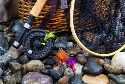 Closeup of fishing fly reel, landing net, creel and assorted flies on wet river bed stones
