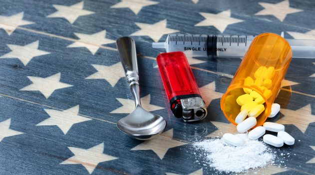 Front view of opioid pain killer tablets with spoon, lighter and syringe on rustic USA flag in background for drug addiction concept in America  