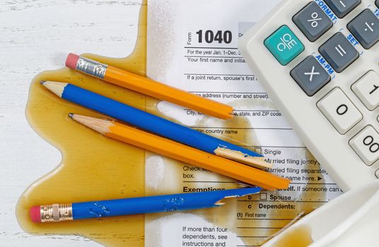 Top view U.S. Individual tax form 1040, coffee spilled out of paper cup,  with partial old calculator and broken pencils on wooden desktop.