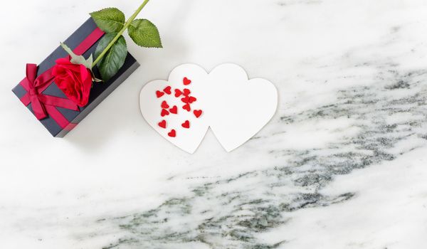 Single red rose on dark gift box with heart shaped card and candy on marble stone background in flat lay view 