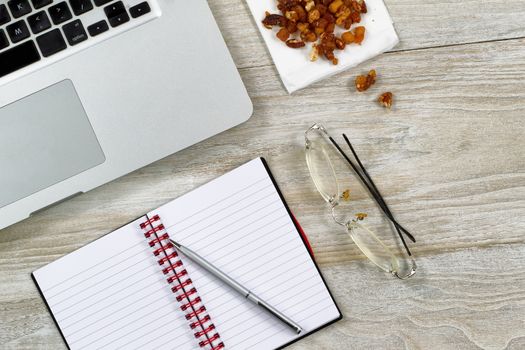 Top view of business office consisting of computer, notepad, pen, reading glasses, and snack food on rustic white wooden desktop 
