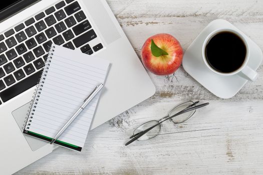 Top view of desktop with partial laptop, apple, coffee, reading glasses, and pen. 