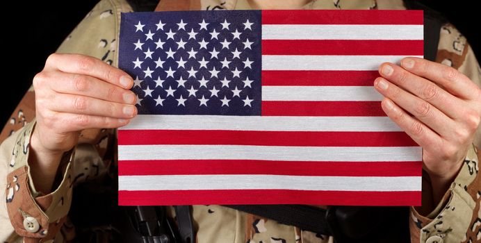 Close up horizontal image of United States of America flag with armed male soldier holding it while on black background. 