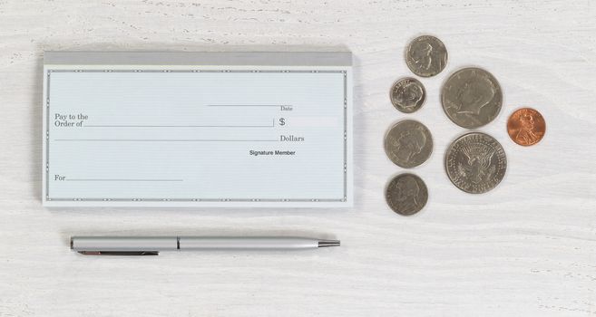 Close up of blank checkbook, silver pen and coins on white wooden desktop. Layout in horizontal format.