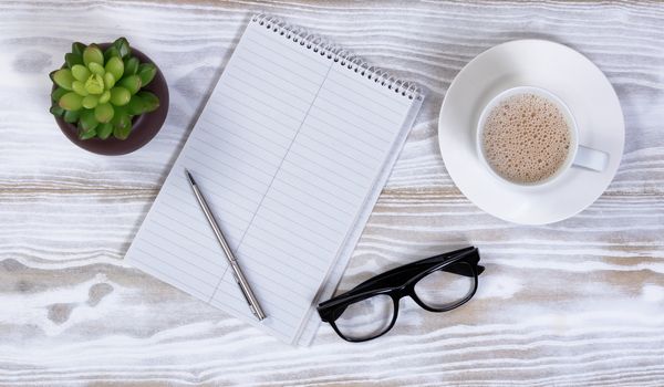 Notebook and pen with reading glasses, plant and creamy coffee on rustic white table