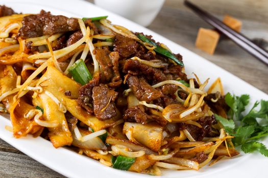 Close up front view of freshly cooked spicy beef, bean sprouts, onion, and tofu with parsley as garnish and chopsticks in holder on rustic wood. 