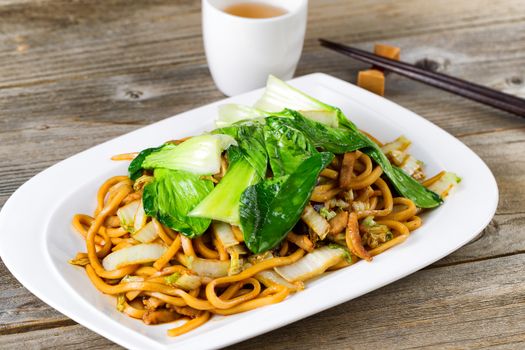 Close up front view of spicy cooked noodles, onion, bok choy, and chicken slices. Chopsticks and tea in background on rustic wood. Selective focus on middle of dish. 