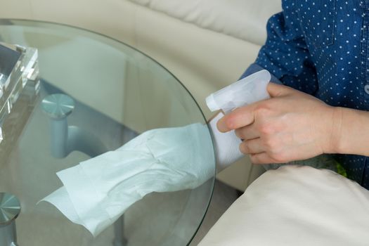 Horizontal photo female hands wiping underneath glass round end table with spraying cleaning solution bottle and paper towels in hand with sofa and partial lamp in background 