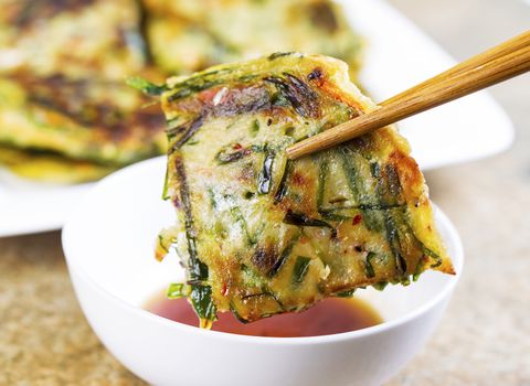 Horizontal photo of Korean Green Onion Pancake being held with chopsticks while be dipped into small cup of sauce in front of plate filled with more pancakes