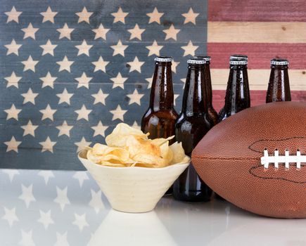 Football, potato chips, cold beer in bottles on white glass table with United States flag painted on rustic wood. Layout in horizontal format with copy space. 
