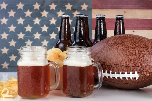 Pint glasses of beer, football, bowl of potato chips, cold beer in bottles on white glass table with United States flag painted on rustic wood. Layout in horizontal format. 