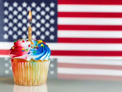 Close up of a red, white, and blue frosted cupcake with burning candle. Fourth of July concept with United States of American flag in background.