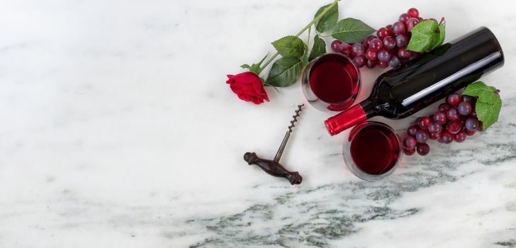 Overhead view of Red wine bottle with grapes, drinking glasses, corkscrew and rose on natural marble stone