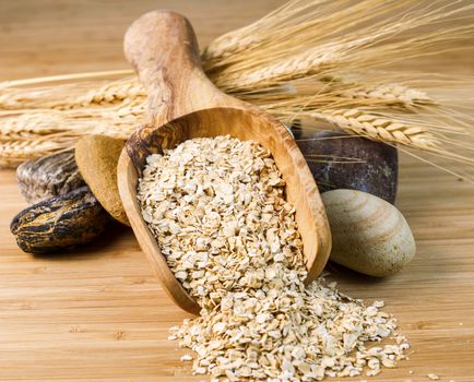 Horizontal photo of rolled oats in wooden spoon with round stones and wheat stalks on natural bamboo wood  