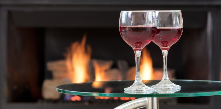 Two glasses of red wine on top of glass table with glowing fireplace in background 