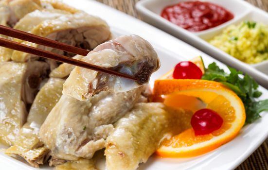 Close up front view of a succulent sliced chicken, selective focus on single piece in chopsticks, with dipping sauces and garnishes. 