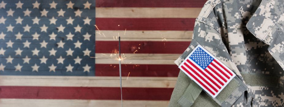 Military uniform with United States of America patch flag and burning sparkler in background on rustic wooden flag. July 4th holiday concept. 