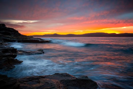 A fiery red sky as the sunrise light colours the clouds in shades of red and orange.  Waves move in and around the rocky shore line, reflecting the sky  and shimmering in the early morning