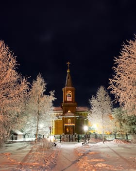 The building of the Orthodox Church at night surrounded by trees in the snow illuminated by lanterns.