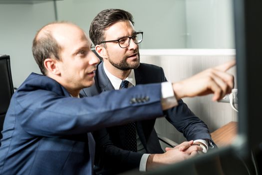 Image of two thoughtful businessmen looking at data on multiple computer screens, solving business issue at business meeting in modern corporate office. Business success concept.