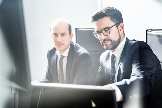 Image of two thoughtful businessmen looking at data on multiple computer screens, solving business issue at business meeting in modern corporate office. Business success concept.