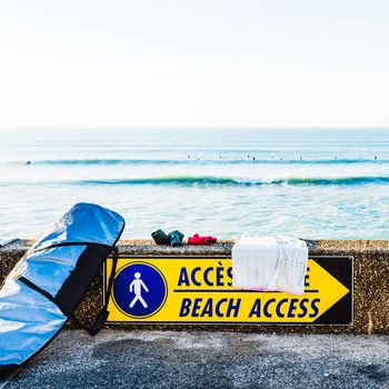 Surfboard in its bag next to a beach access sign, ocean in the background with silhouettes of surfers, in Bidart, France.