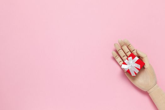 Trendy wooden mannequin hand holds small red gift box on pastel pink background, copy space, flat lay. March 8th, February 14th, birthday, Valentine's, Mother's, Women's day celebration concept.