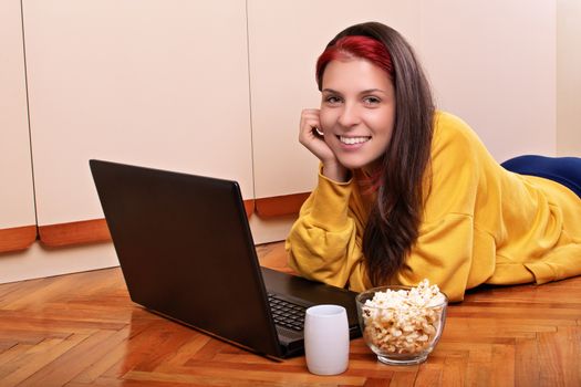 Beautiful smiling young girl in casual clothes lying on the floor of her bedroom, ready to watch a movie on her laptop with a bowl of popcorn and a cup of coffee or tea. Leisure concept.