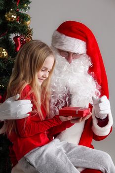 Portrait of smiling little girl sitting on santa claus knees near christmas tree and getting a gift