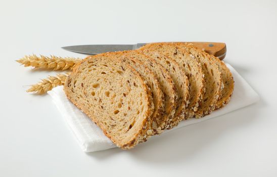 Sliced loaf of whole grain bread on white napkin, wheat ears and kitchen knife next to it