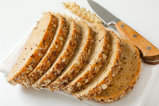 Sliced loaf of whole grain bread on white napkin, wheat ears and kitchen knife next to it