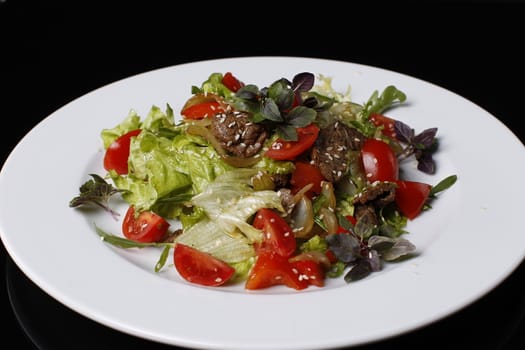 On a white plate salad of tomatoes, fried meat, onions, lettuce, cabbage strewed with sesame seeds, basil, on a black background.Dietary food