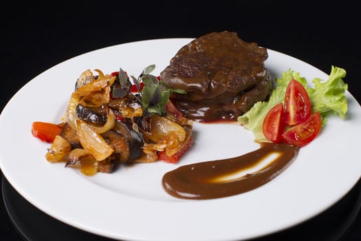 On a white plate fried meat with gravy, stewed vegetables, lettuce leaves, sauce, basil and tomato cut.Beef in sauce.