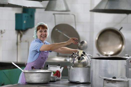 Woman cook in an industrial kitchen pours a drink into the kettle with a large scoop and smiles.The cook of the restaurant