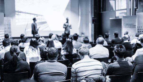 Male public speaker giving a talk at business meeting conference. Rear view of unrecognized people sitting in audience in the conference hall. Business and entrepreneurship concept. Blue toned.