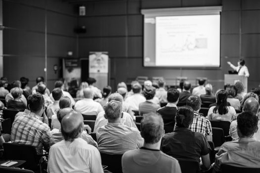 Business and entrepreneurship symposium. Speaker giving a talk at business meeting. Audience in conference hall. Rear view of unrecognized participant in audience. Black and white.