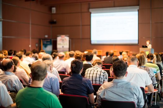 Business and entrepreneurship symposium. Speaker giving a talk at business meeting. Audience in conference hall. Rear view of unrecognized participant in audience.