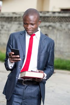 Young student holding a book in his left hand and manipulating a cell phone with the other hand