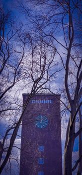 Red brick clock tower against clear blue sky and white clouds, seen through tree branches. Clock tower of the central train station in Oberhausen, Germany.