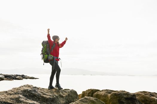 Woman with backpack and arms raised  enjoying the beautiful morning view of the coast