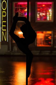 Dancing elegant young girl hanging in a dance pole in front of windows with red light and open sign