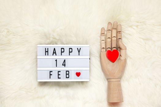 Trendy wooden human hand model holds red heart, lightbox with inscription Happy 14 FEB that means Valentine's Day on white fur carpet. Flat lay. February 14th celebration concept. Horizontal.