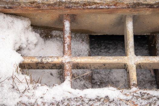 A storm drain on the side of a country road leading to a bridge has a dusting of snow in the winter time. The metal grating over top is gradually rusting, exposed to the elements.