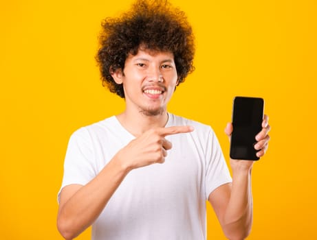 Happy asian handsome young man with curly hair holding blank screen smart phone and pointing finger isolate on yellow background