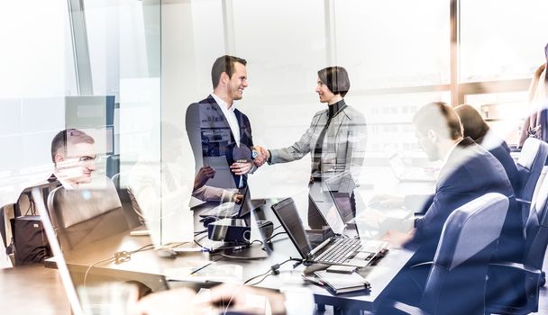 Sealing a deal. Business people shaking hands, finishing up meeting in corporate office. Businessmen working on laptop seen in glass reflection. Business and entrepreneurship concept.