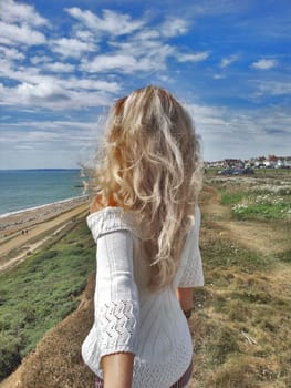 Follow me. Blonde girl standing back on the edge cliff, against blue sky background