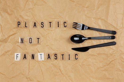 Disposable black appliances fork, spoon, knife, phrase from letters on transparent base Plastic not fantastic on brown crumpled craft paper. Eco, zero waste concept. Flat lay, top view. Horizontal.