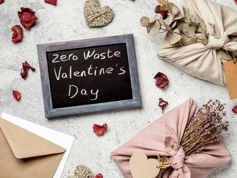 Zero waste Valentine's Day concept. Eco-friendly gift cloth wrapping in Furoshiki style and chalkboard with Zero Waste Valentine's Day letters on gray textured background. Top view or flat lay