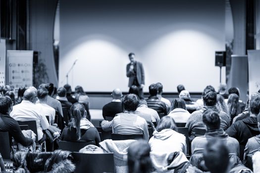 Male public speaker giving a talk at business meeting conference. Rear view of unrecognized people sitting in audience in the conference hall. Business and entrepreneurship concept. Blue toned.