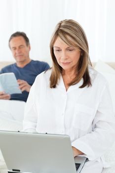 Woman looking at her laptop while her husband is reading on the bed
